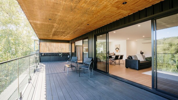 A modern outdoor deck with wooden flooring, metal chairs, and a glass door leading to an interior dining area with a sofa and table. - Tree Top Haven ITAS - Hosted by L'Abode (South Launceston)