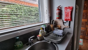 A kitchen with a sink, window blinds, a fire extinguisher, a fire blanket, and a wooden cutting board.