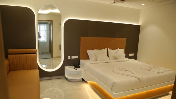 A modern hotel room with a large bed, a bedside table, a mirror, and a seating area. - SRIPADAM GRAND (Chilakaluripet)