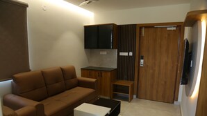 A compact living space with a brown sofa, a small white table, a TV on a stand, wooden cabinets, and a door with a vertical panel design. - SRIPADAM GRAND (Chilakaluripet)