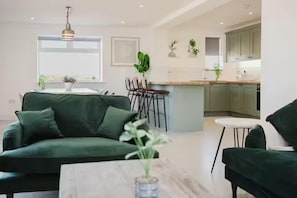 A modern living room with a green sofa, a coffee table, and a dining area with chairs. A kitchen is visible in the background with light-colored cabinets and a central island. - Gatekeepers Cottage - 4 Bed Rural Escape (Cropthorne)