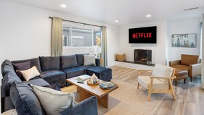 A modern living room with a sectional sofa, a wooden coffee table, a fireplace, a TV mounted on the wall, and a comfortable armchair. - Steps to Sand! 2 Homes in 1 – Sleeps 17, Outdoor Space, Walk to Shops & Dining (Newport Beach)