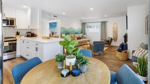 A modern kitchen with white cabinets, a built-in microwave, and a stove. There is a dining area with a round wooden table and blue chairs. - Steps to Sand! 2 Homes in 1 – Sleeps 17, Outdoor Space, Walk to Shops & Dining (Newport Beach)