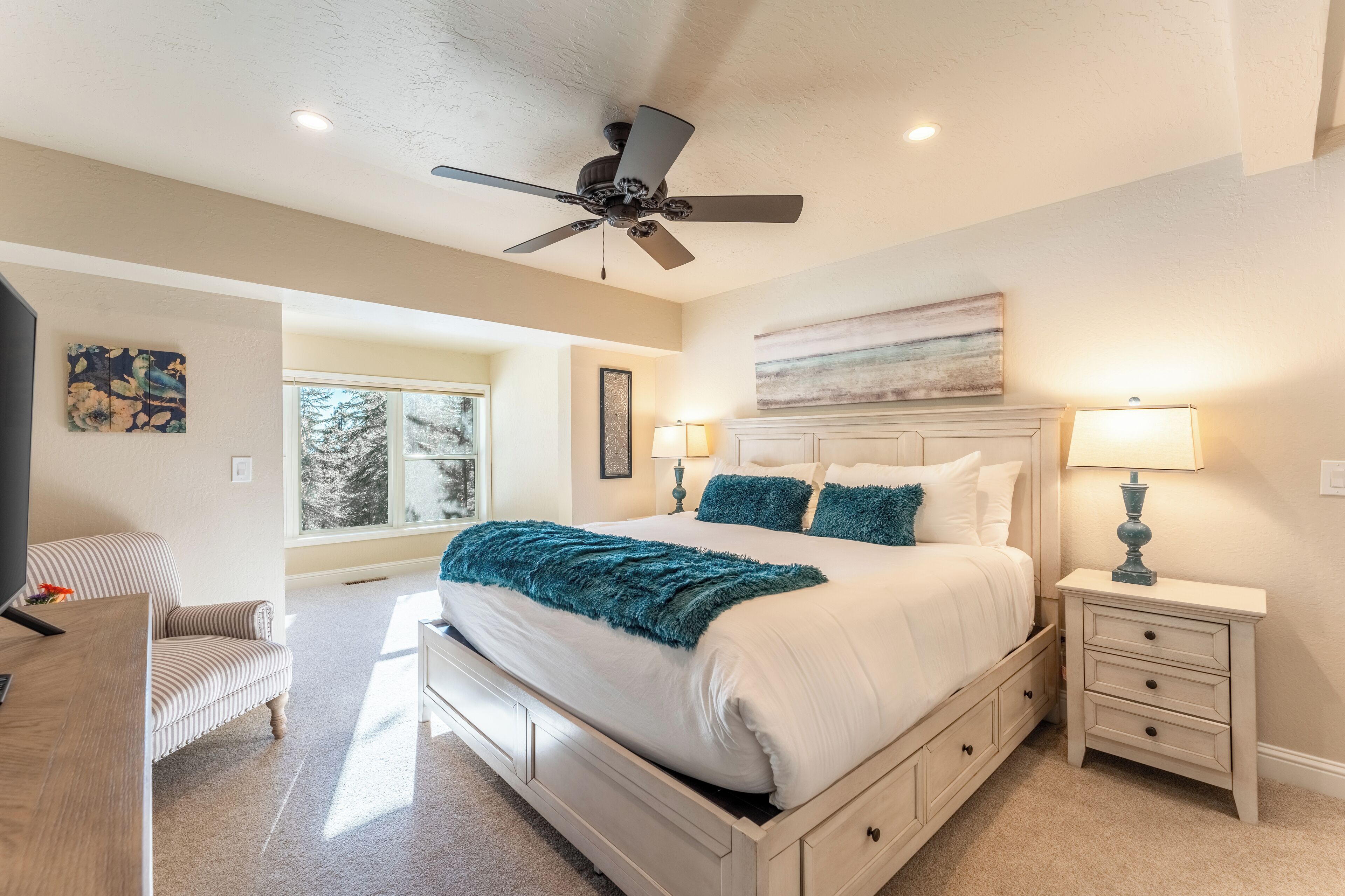 A bedroom with a large bed, two bedside tables with lamps, a ceiling fan, and a window with a view of trees.