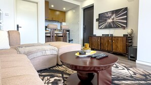 A modern living room with a sectional sofa, a round coffee table, and a kitchen in the background. - Kilimani Bliss,Alba gardens studio apartment with a gym, infinity swimming pool (Nairobi)