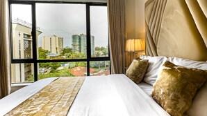 A hotel room with a large window, a bed with white linens and gold pillows, a headboard with vertical ridges, and a lamp on a bedside table. - Kilimani Bliss,Alba gardens studio apartment with a gym, infinity swimming pool (Nairobi)