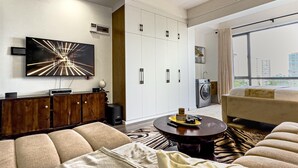 A modern living room with a sofa, a coffee table, and a large painting. There is a wardrobe with a mirror and a window with a view of buildings. - Kilimani Bliss,Alba gardens studio apartment with a gym, infinity swimming pool (Nairobi)