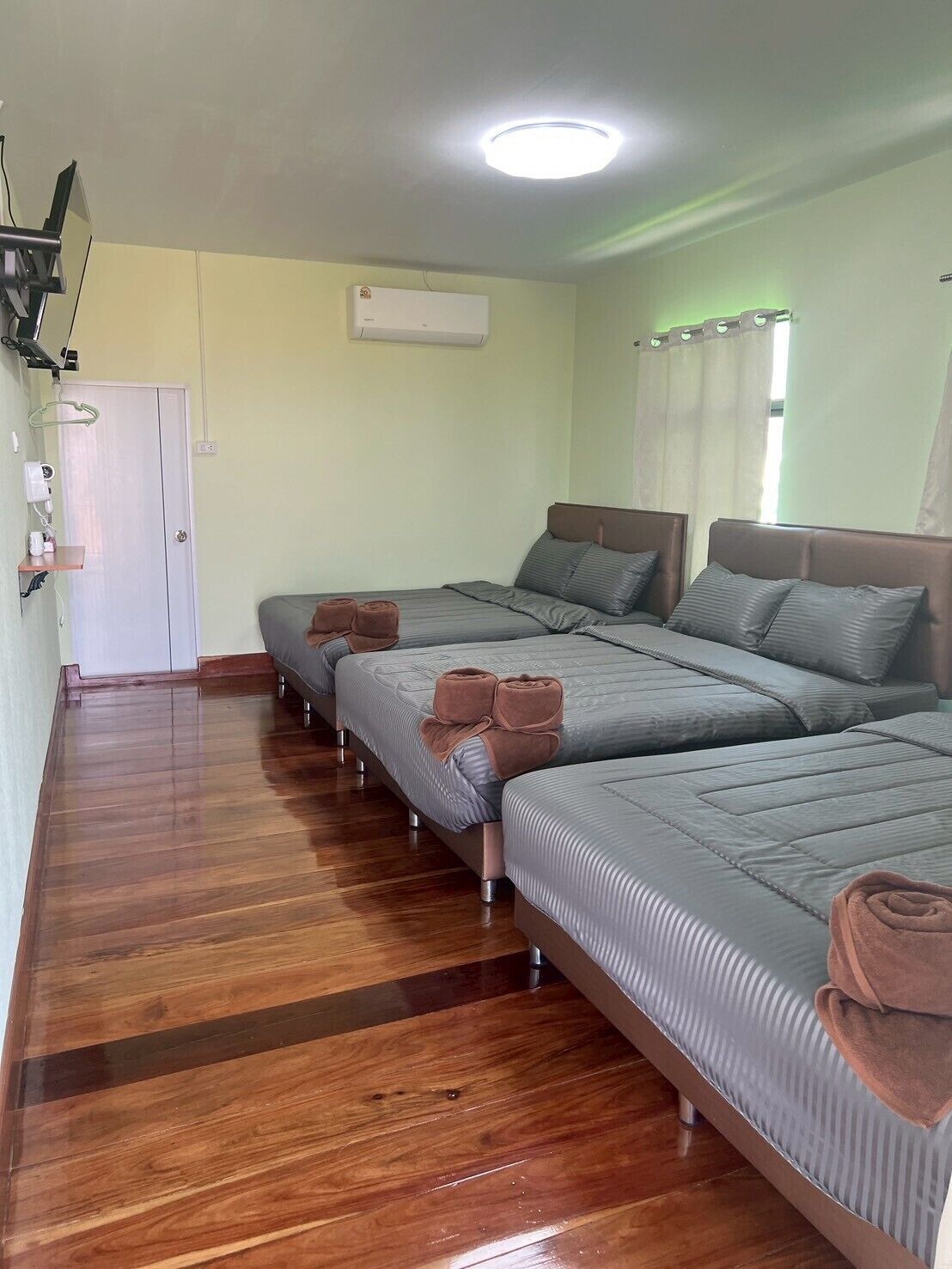 Een hotelkamer met twee bedden, een houten vloer, een witte deur, een airconditioning en een raam met gordijnen.