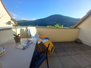Uma varanda com uma mesa, cadeiras, um vaso de planta e vista para uma montanha.