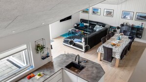 A modern kitchen with a central island, a dining area with a table and chairs, a black leather sofa, and a view of the outdoors through a large window.