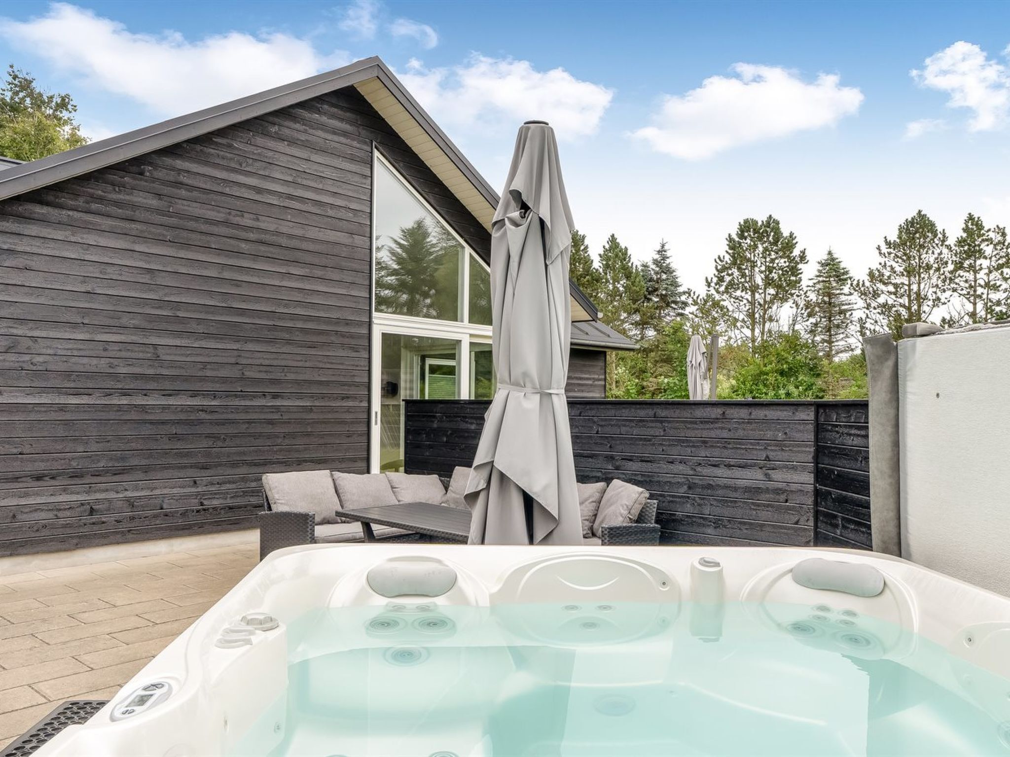 A modern outdoor hot tub area with a wooden deck, a seating area, and a large window.