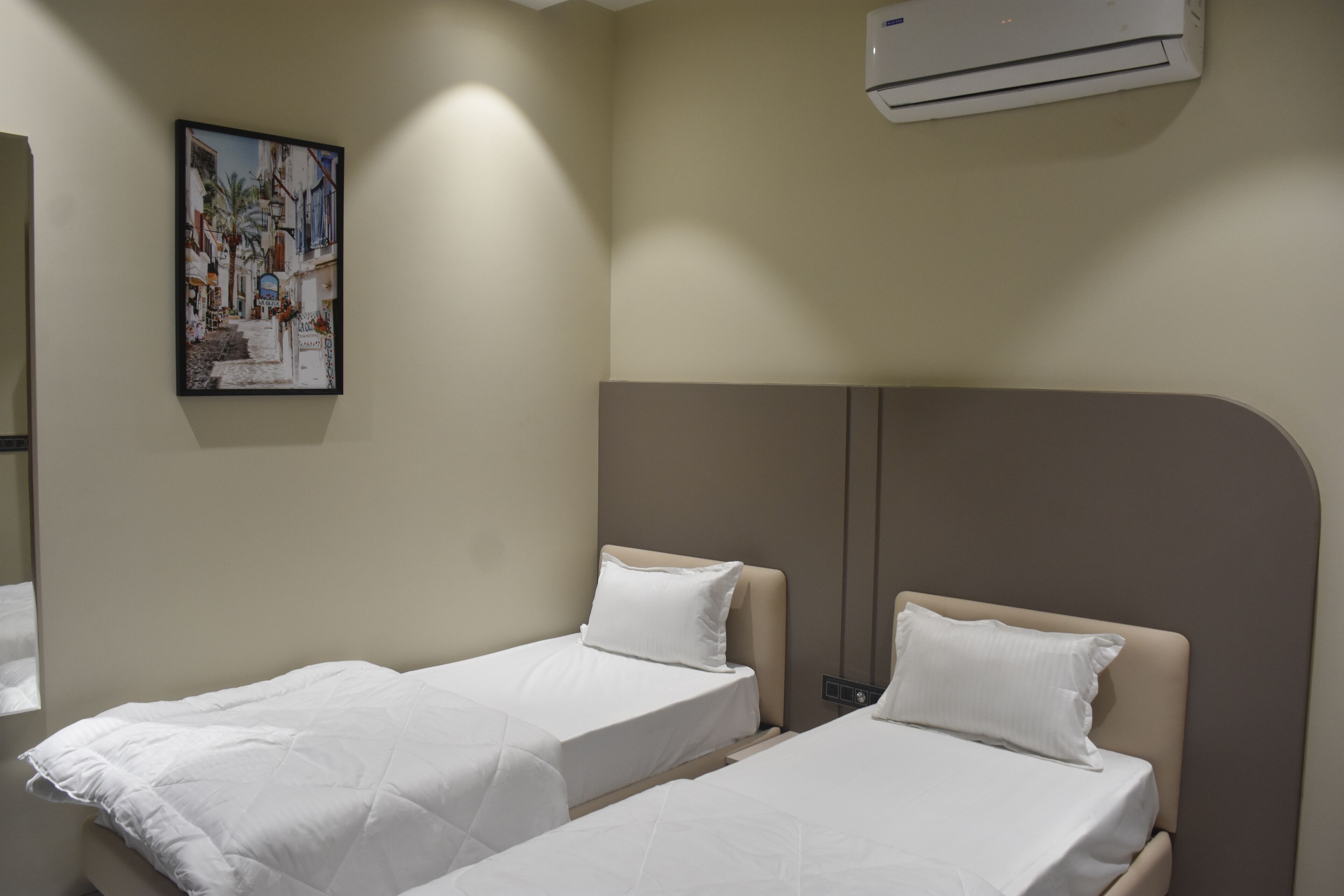 A hotel room with two single beds, a painting on the wall, and an air conditioning unit.