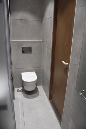 A small, modern bathroom with a white toilet, a wooden door, and grey tiled walls. - Hotel Elite (Bhachau)