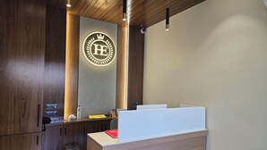 A reception desk with a wooden finish, a logo on the wall, and a ceiling with wooden beams and pendant lights. - Hotel Elite (Bhachau)