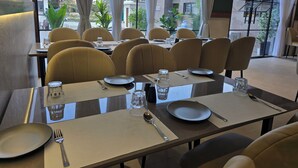 A dining area with tables set for four, each with plates, cutlery, and glasses. The room has large windows with curtains, allowing natural light and views of the outside. - Hotel Elite (Bhachau)