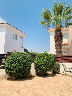 A residential building with a tiled patio, two well-maintained bushes, a palm tree, and a chair.