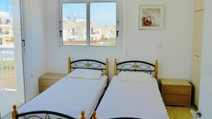 A room with two single beds, a window with a view of buildings, a small bedside table, and a wardrobe.
