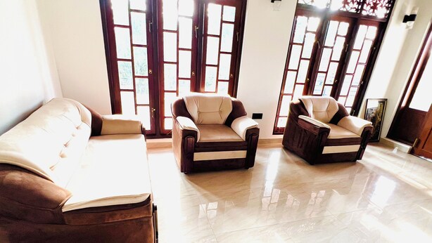 A spacious living room with a brown sofa and two armchairs, large windows with decorative glass panels, and a tiled floor. - 5-bedroom villa in wonderful Negombo, 10 mins away from Airport & Beach. (Negombo)