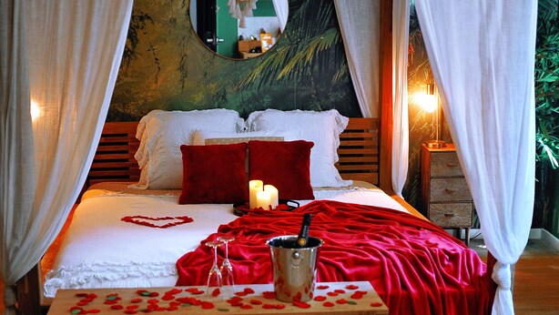 Un dormitorio con cama, ropa de cama roja, una mesita de noche con velas y un espejo en la pared.