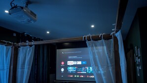 Un cine en casa con pantalla proyectora mostrando la interfaz de Netflix, un refrigerador pequeño y cortinas.