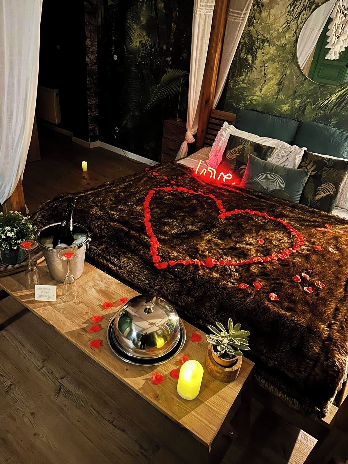 Une chambre confortable, décorée d’un motif en forme de cœur sur le lit, avec une table de chevet en bois sur laquelle est posée une bougie, et une fenêtre donnant sur les arbres.