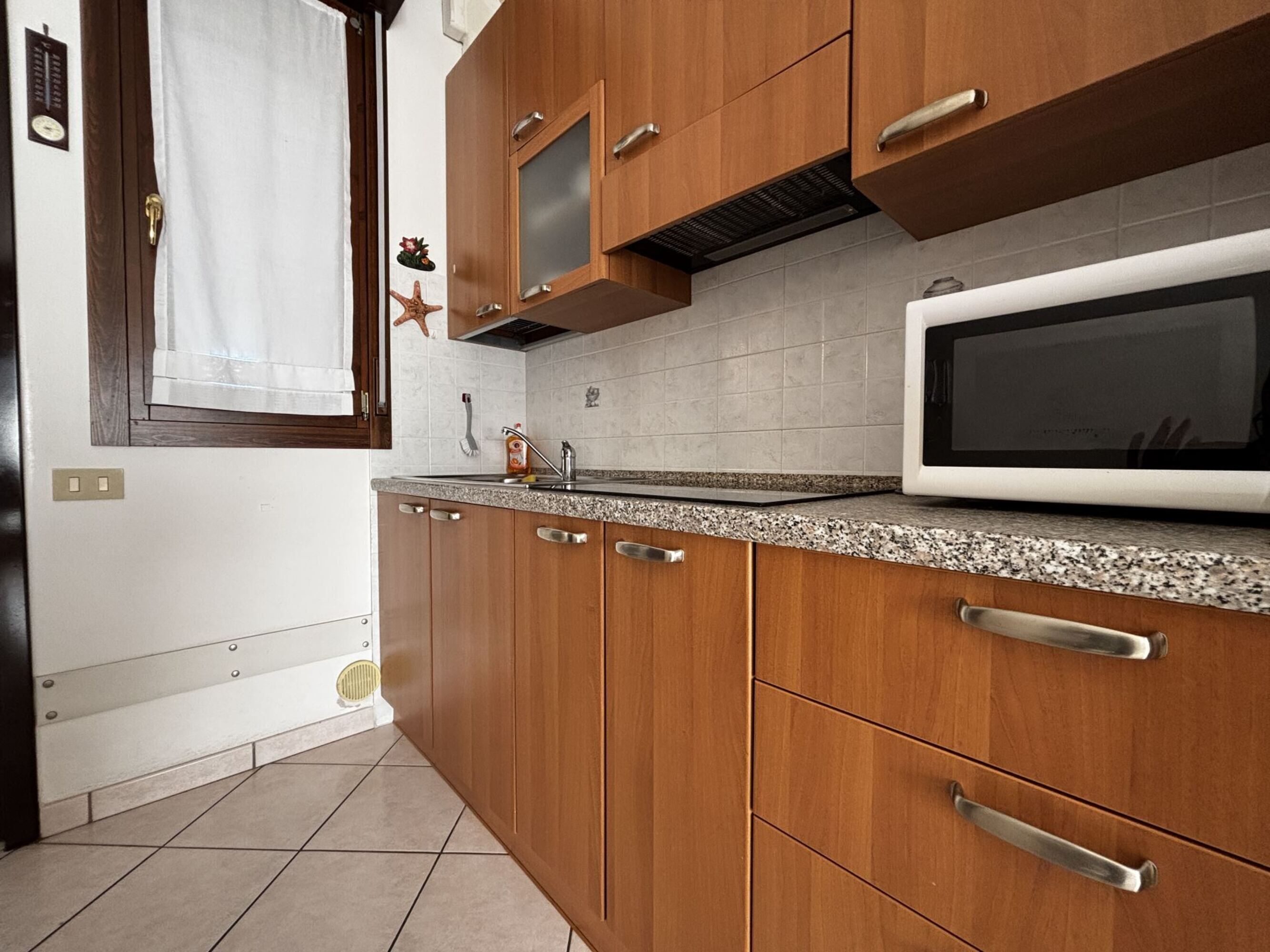 A kitchen with wooden cabinets, a granite countertop, a microwave, and a window with a curtain.