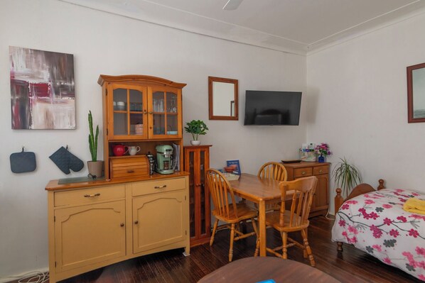 A cozy room with a wooden cabinet, a dining table with chairs, a bed with a floral bedspread, a television, and a potted plant. - Unit 1 - A welcoming and cosy unit, near Wanda beach and Cronulla Golf Course (Cronulla)