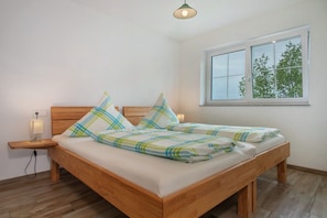A double bed with a wooden frame, a nightstand with a lamp, and a window with a view of trees.