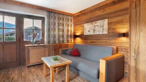 Een gezellige kamer met houten inrichting, een grijze bank, een klein houten tafeltje en een raam met gordijnen.