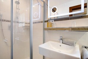 A bathroom with a glass shower enclosure, a white sink, and a mirror with a round light.