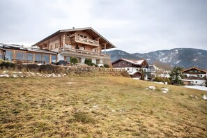 A large wooden chalet with a balcony, surrounded by a hilly landscape and mountains in the background. - Studio 'Panorama - Apartment 3' with Mountain View, Private Balcony and Shared Sauna (Reit im Winkl)