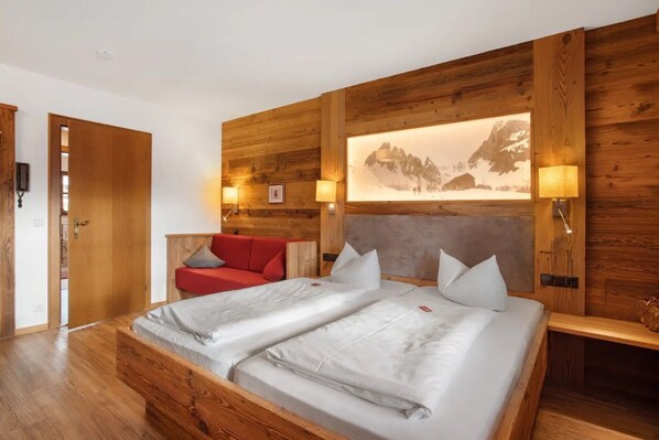 A hotel room with a wooden headboard, a bed with white linens, a red sofa, and a mountain landscape painting on the wall. - Studio 'Panorama - Apartment 3' with Mountain View, Private Balcony and Shared Sauna (Reit im Winkl)