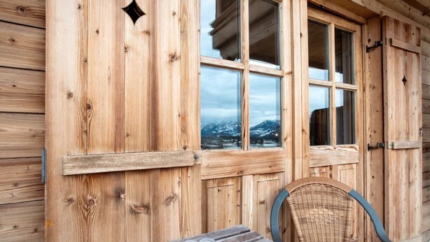 A wooden cabin with a table and chairs, a window with a view of snow-capped mountains, and a door with a handle. - Studio 'Gästehaus Panorama - Apartment 5' with Mountain View, Private Balcony & Shared Sauna (Reit im Winkl)