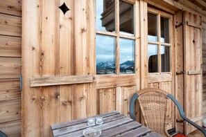 Een houten chalet met een tafel en stoelen, een raam met uitzicht op besneeuwde bergen, en een deur met een klink.
