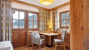 A cozy room with wooden floors, a round wooden table, two chairs, a lamp, and large windows with curtains. - Studio 'Gästehaus Panorama - Apartment 5' with Mountain View, Private Balcony & Shared Sauna (Reit im Winkl)