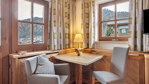A cozy room with a wooden table, two grey chairs, a lamp, and large windows with curtains. - Studio 'Gästehaus Panorama - Apartment 5' with Mountain View, Private Balcony & Shared Sauna (Reit im Winkl)