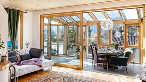 Ein modernes Interieur mit Holzbalkendecke, einer Couch mit Kissen, einem Esstisch mit Stühlen und großen Glasüren, die zu einem Außenbereich führen.
