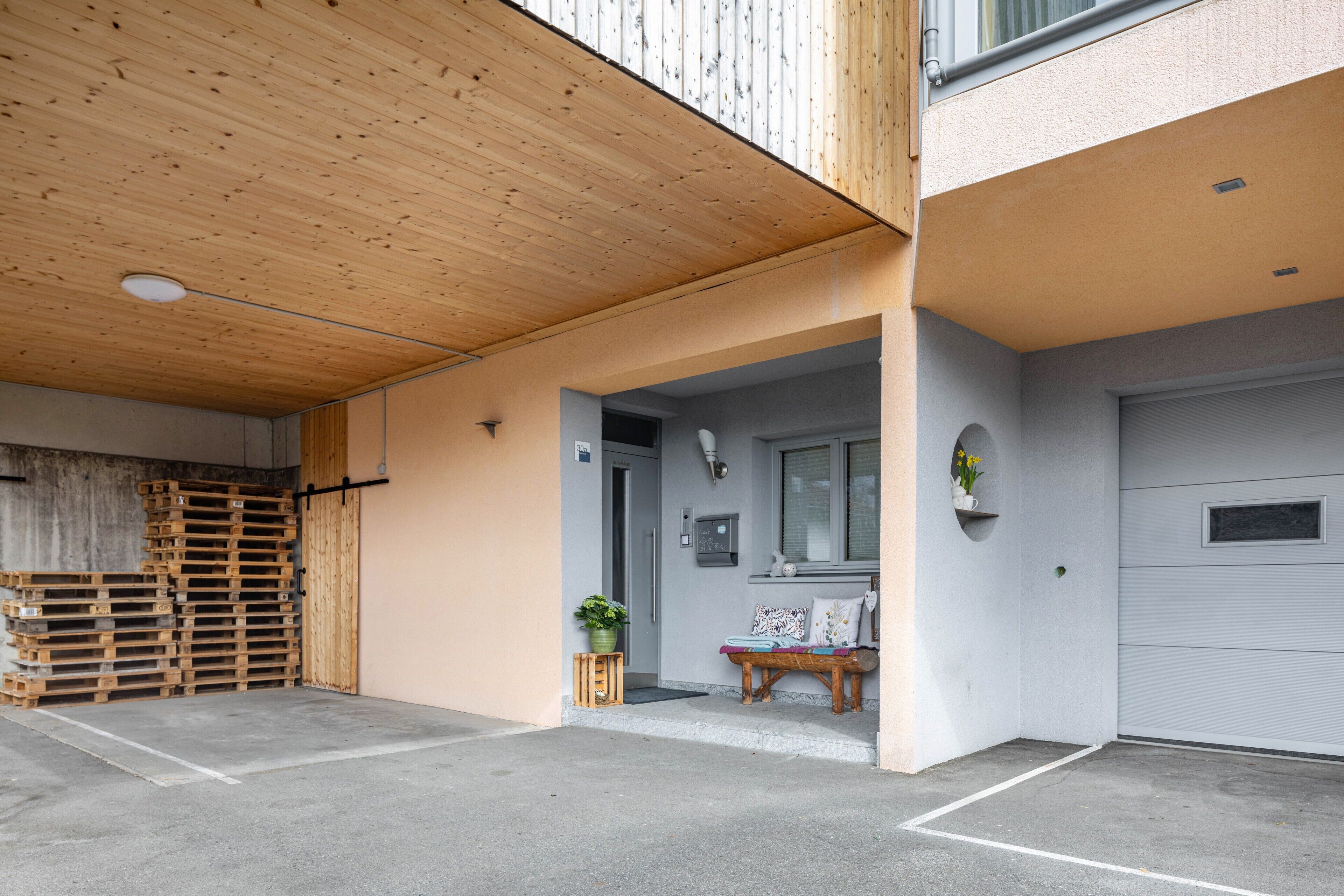 Un immeuble résidentiel doté d’un plafond en bois, d’une porte de garage, d’un petit banc avec des coussins et d’une plante en pot.