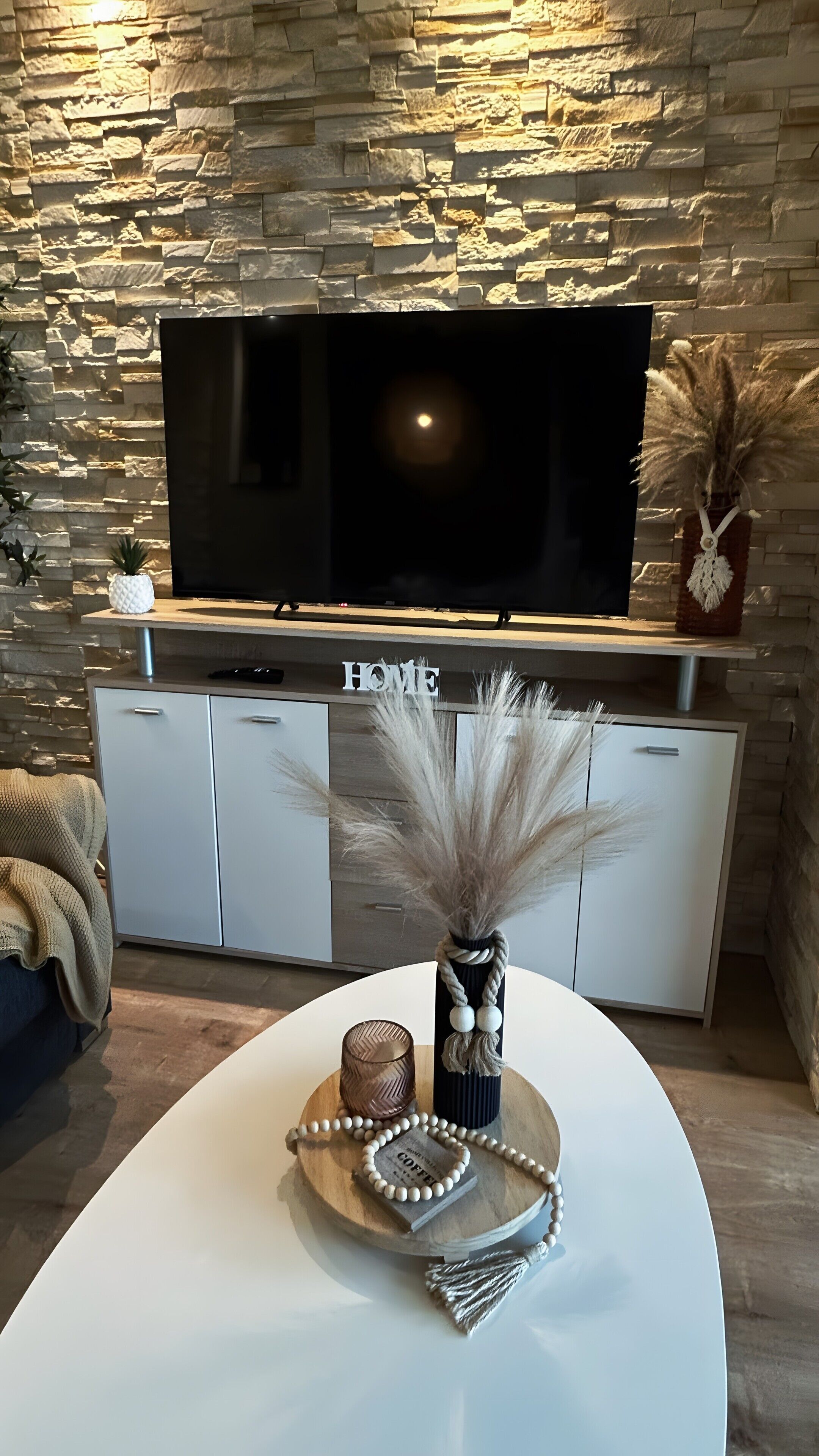 Een moderne woonkamer met een stenen muur, een flatscreen televisie, een witte kast en een decoratieve middelpunt op tafel.