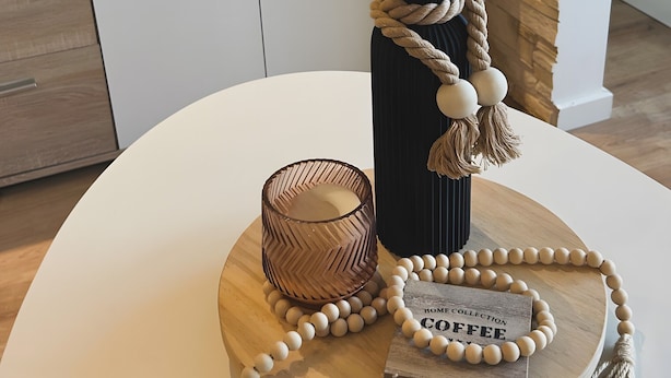 A round table with a vase of pampas grass, a candle, a beaded necklace, and a wooden coaster.