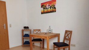 Un área de comedor con una mesa de madera, dos sillas, un estante con sillas plegables y un cuadro en la pared.