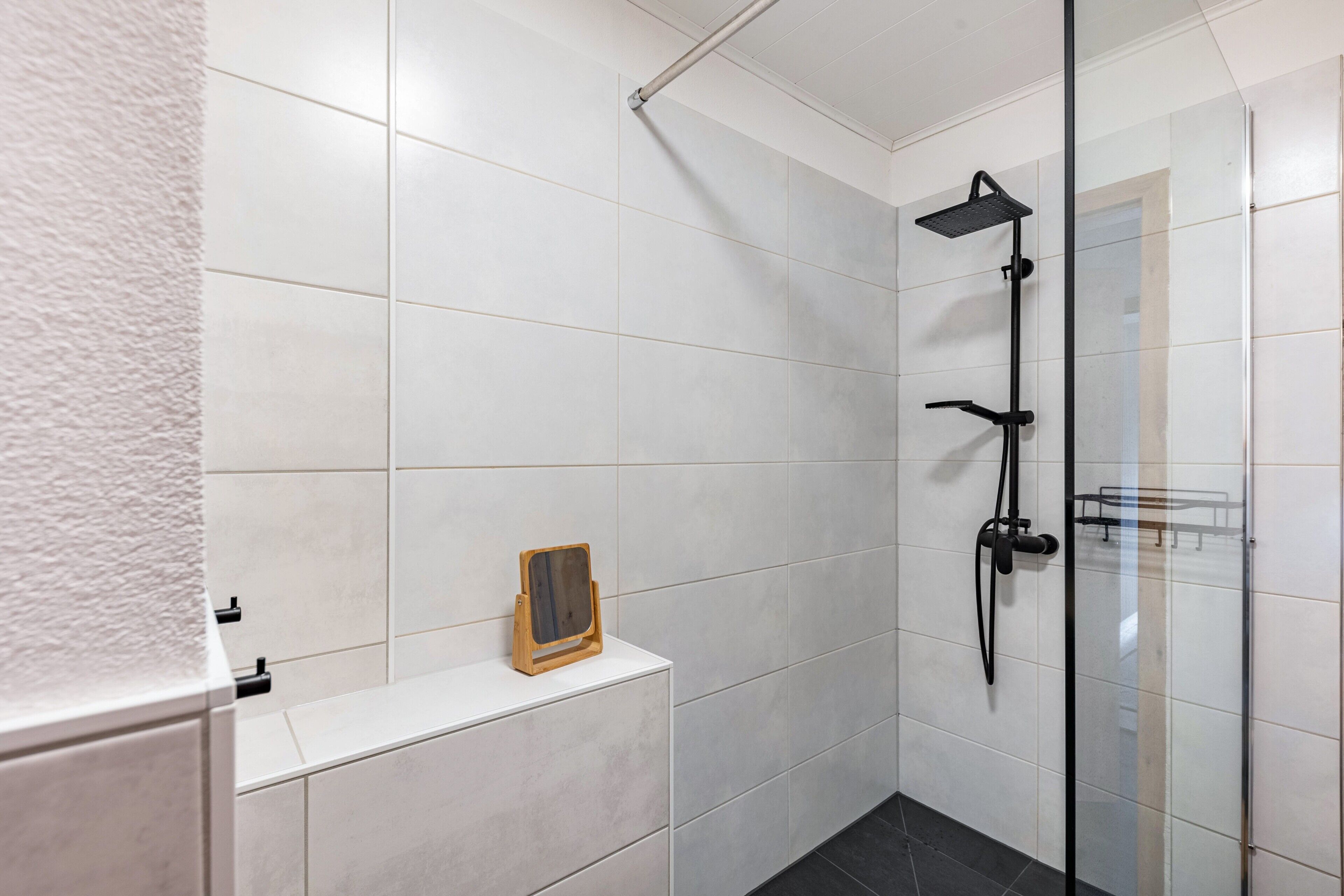Un baño moderno con azulejos blancos, cabina de ducha de cristal y un pequeño estante de madera.