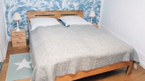 Una cama individual con cabecera de madera, dos lámparas de noche, una manta gris, un tapete con estampado de estrellas y una mesita de noche con una lámpara.