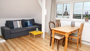 A room with a grey sofa, wooden dining table, chairs, a yellow side table, and a window with a view of a cityscape. - Apartment 'Mit Panoramablick' with Wi-Fi (Flensburg)