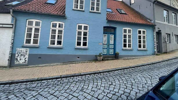 A two-story building with a blue facade, a red-tiled roof, and a cobblestone street in front. - Apartment 'Mit Panoramablick' with Wi-Fi (Flensburg)