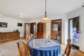 Uma sala de jantar com uma mesa redonda, cadeiras, um candelabro, armários de madeira e vista através de uma porta de vidro.