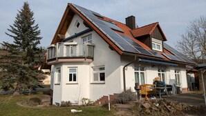 Una casa de dos pisos con techo inclinado, paneles solares, un balcón y un pequeño jardín.