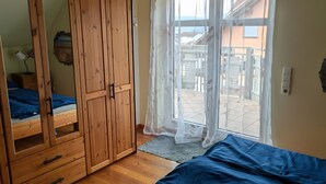 Un dormitorio con un armario de madera, una cama con cubrecama azul, un ventilador de techo y vistas a través de una ventana con cortinas de encaje.