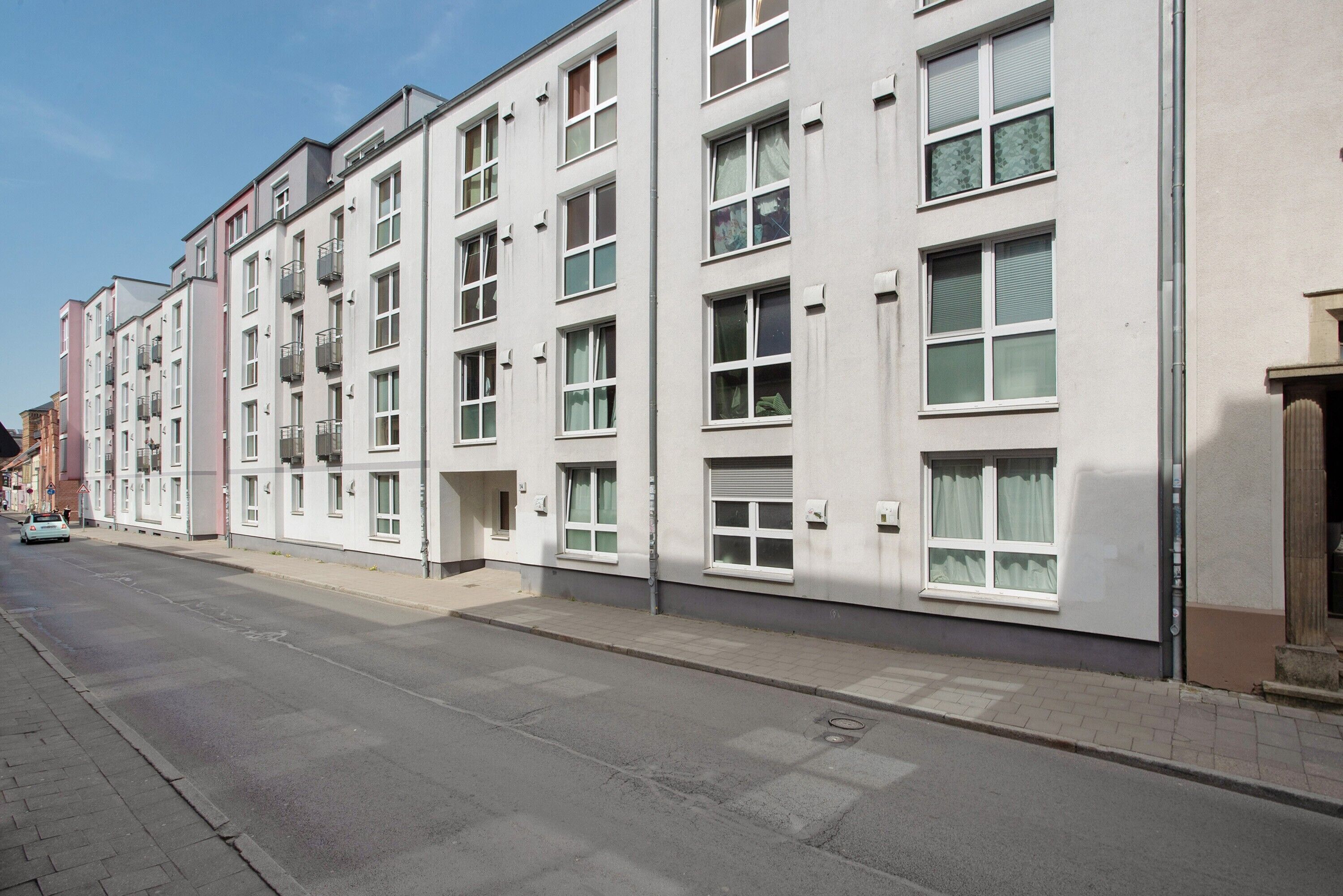 A row of multi-story residential buildings with balconies, a paved street, and a clear sky.