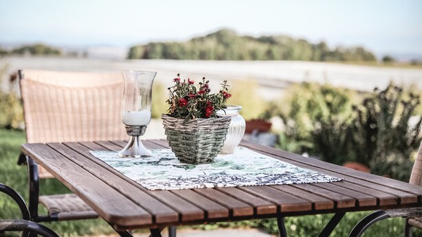 Een houten tafel met een placemat, een rieten mand met bloemen, een witte vaas en een rieten stoel.